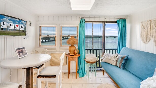 A cozy room with a blue sofa, a white table with chairs, a small wooden table with a lamp, and a view of the sea through large windows. - Bliss on the Bay by Seaside Vacations & Sales (Chincoteague Island)