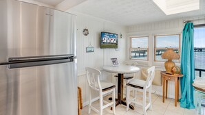 A kitchen with a stainless steel refrigerator, a round dining table with white chairs, and a television mounted on the wall displaying 'LOVE'. - Bliss on the Bay by Seaside Vacations & Sales (Chincoteague Island)