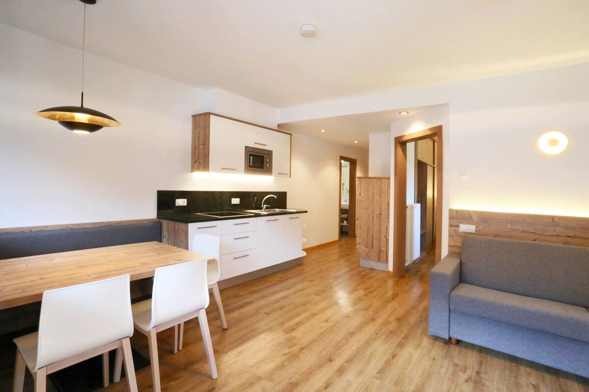 Eine moderne Küche mit einem Holzes Esstisch und Stühlen, einer eingebauten Mikrowelle und einem grauen Sofa in einem hellen, offenen Wohnbereich.