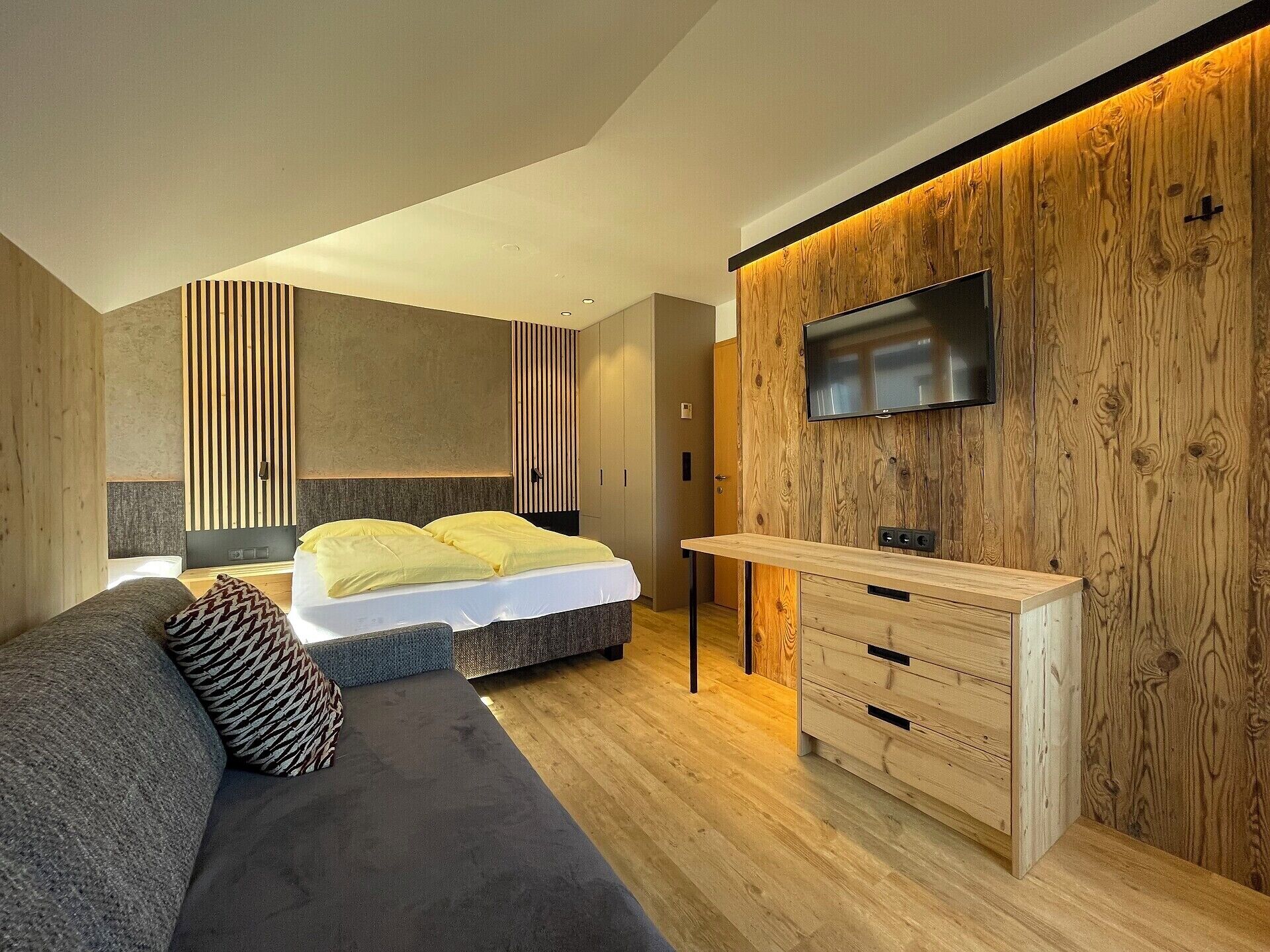 A modern bedroom with a bed, a sofa, a TV mounted on a wooden wall, and a small desk.