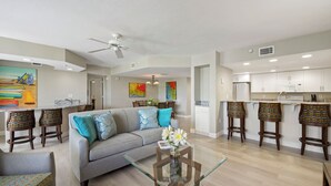 A modern living room with a glass coffee table, a grey sofa, and a kitchen area with white cabinets and a bar stool. - Beach Front - Heated Pool- Partial View- 2BR Gullwing #203 (Fort Myers Beach)