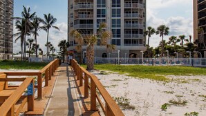 A wooden boardwalk leading to a high-rise building with balconies, surrounded by palm trees and a clear sky. - Beach Front - Heated Pool- Partial View- 2BR Gullwing #203 (Fort Myers Beach)