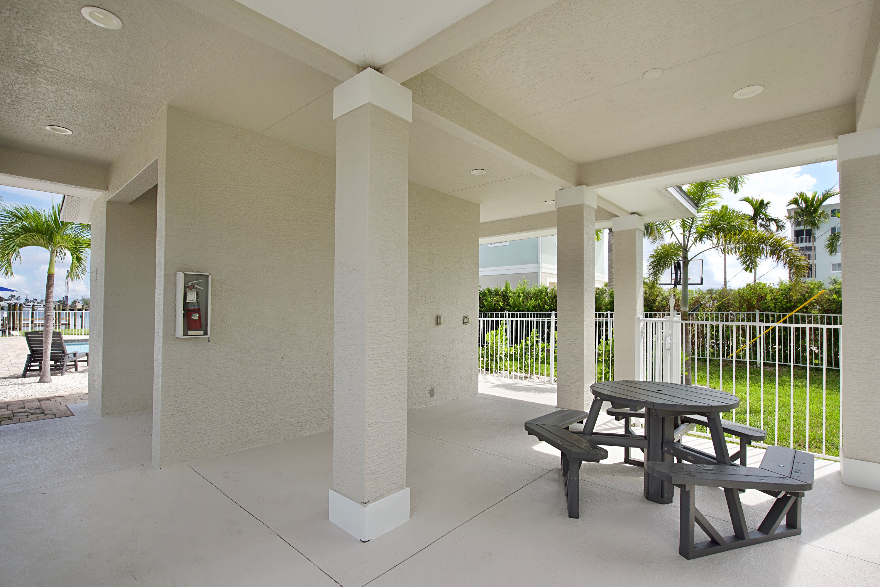 An outdoor patio area with a picnic table and benches, surrounded by white pillars and a white fence.