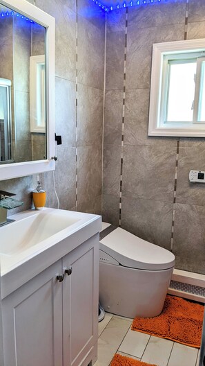 Une salle de bain avec un lavabo blanc, un miroir et des toilettes. Il y a une fenêtre avec des lumières LED bleues au-dessus des toilettes.