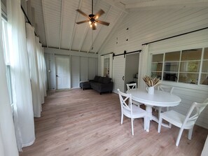 A spacious room with a wooden floor, a ceiling fan, a white dining table with chairs, a dark sofa, and a large window with curtains.