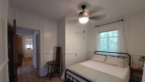 Een slaapkamer met een bed, een plafondventilator, een raam met gordijnen, een nachtkastje en een spiegel.