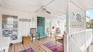 Une véranda avec une pancarte « BIENVENUE », une porte de couleur turquoise, des paniers en osier et une table en bois.
