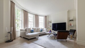 A modern living room with a curved sofa, a coffee table, a TV, and a bookshelf. - Sloane Court East VI by onefinestay (London)