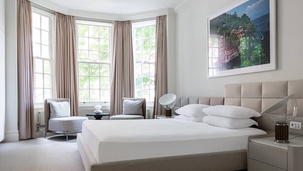 A hotel room with a large bed, a bedside table with a lamp, a chair, and a large window with curtains. - Sloane Court East VI by onefinestay (London)