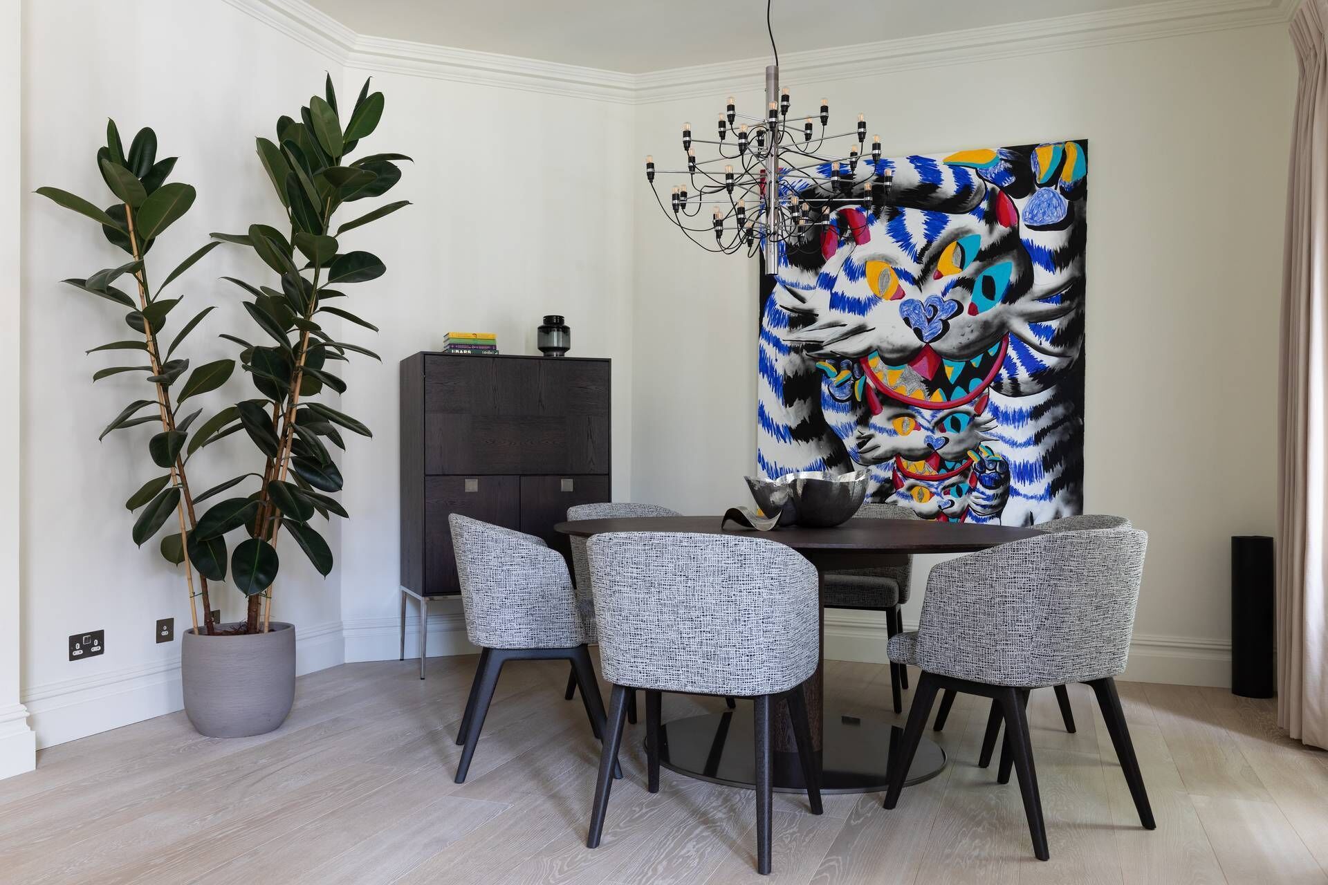 A dining area with a round table, four grey chairs, a black cabinet, a large abstract painting, and two potted plants.