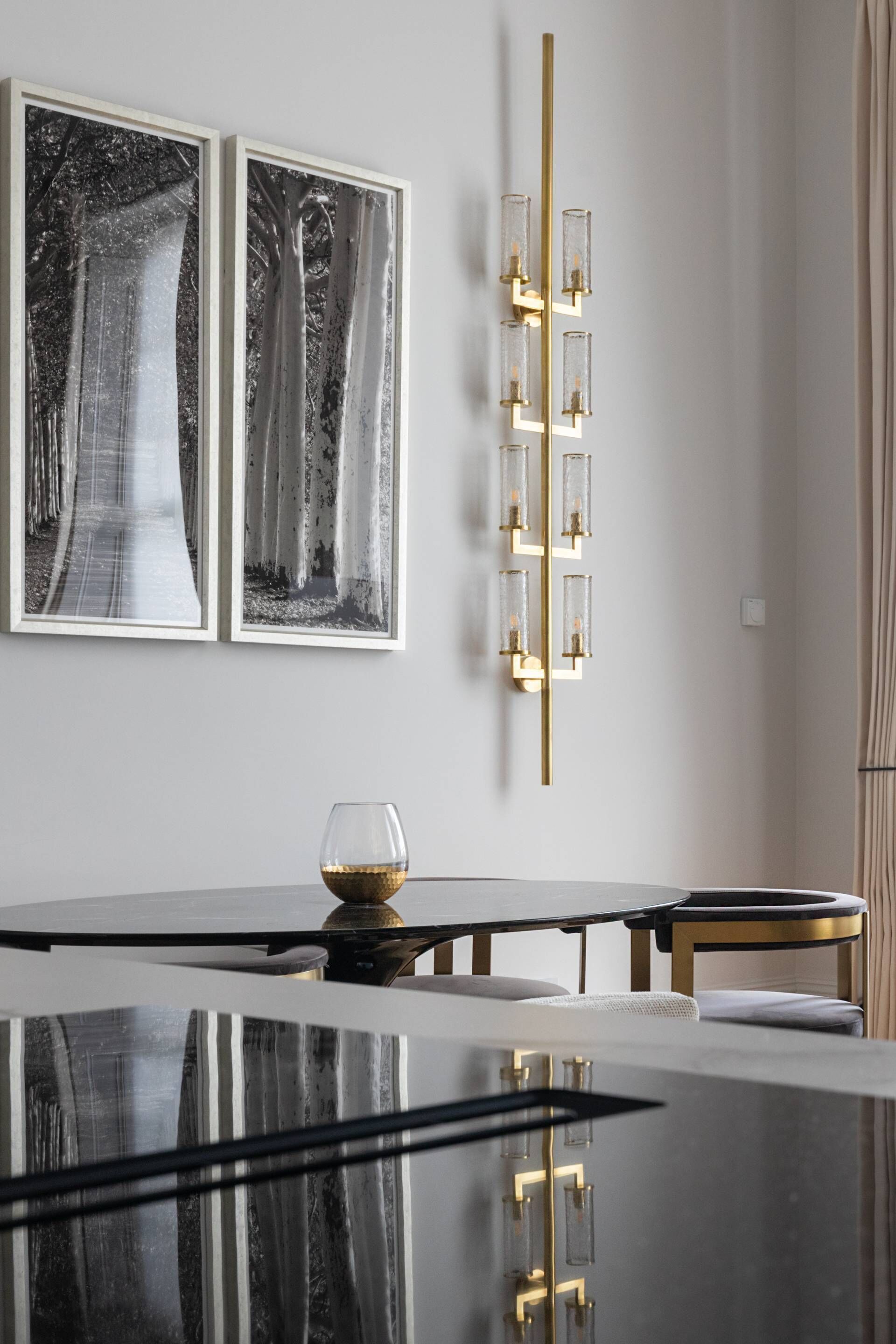 A modern dining area with a reflective black table, two framed black and white photographs on the wall, and a wall-mounted light fixture with multiple glass panels.