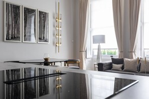 A modern hotel room with a large glass table, a sofa with patterned cushions, and three framed abstract paintings on the wall.
