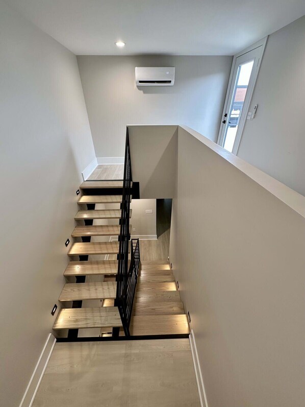 A modern staircase with wooden steps and a black metal railing, leading to a landing with a white wall and an air conditioning unit.