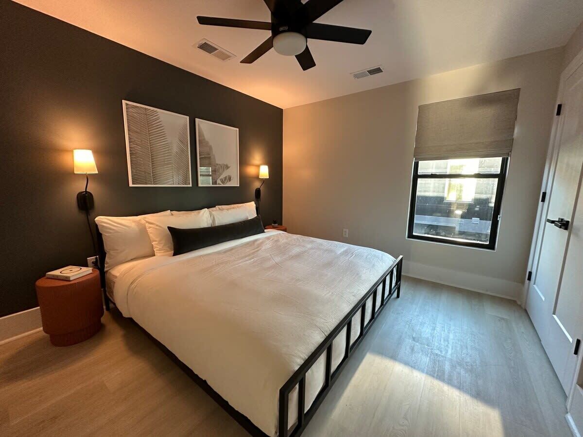 A bedroom with a bed, bedside lamps, a ceiling fan, a window with blinds, and a door.