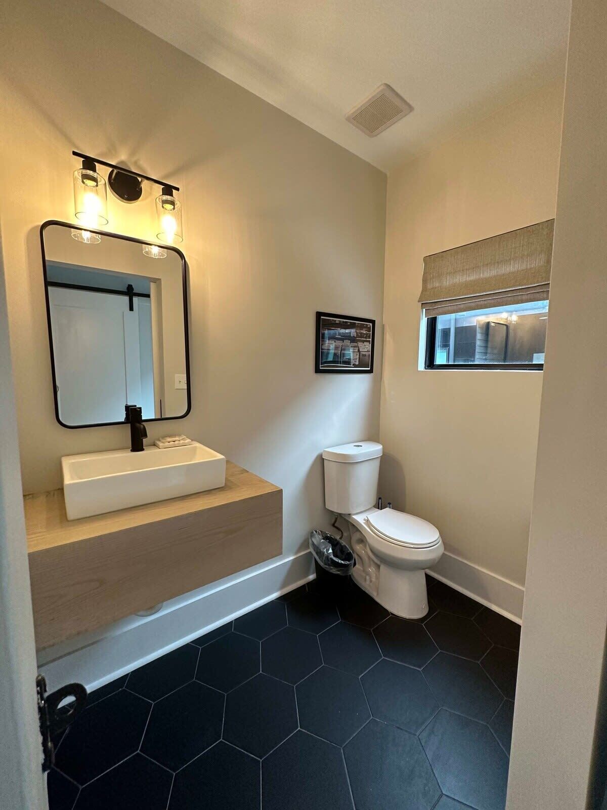 A bathroom with a white sink, black hexagonal tiles, a toilet, and a window with a blind.