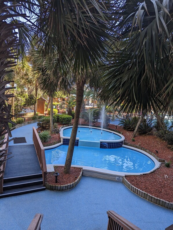 Una piscina circular con una fuente, rodeada de palmeras y una terraza de madera.