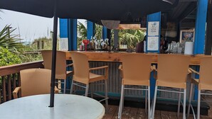 Un espace bar extérieur avec des chaises et une table en osier, une enseigne indiquant « Tito’s Handmade Vodka », et une vue sur l’océan.