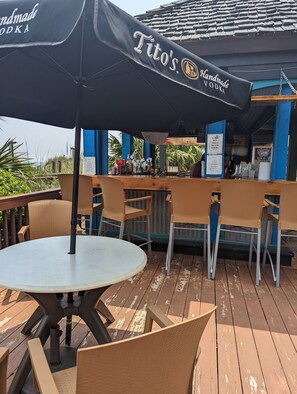 Un área de bar al aire libre con sillas de mimbre y una mesa, un cartel que dice "Tito's Handmade Vodka" y vistas al océano.