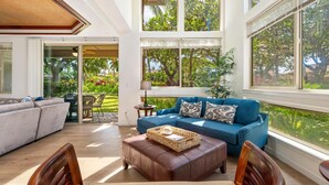 A spacious living area with a blue sofa, leather ottoman, and wooden chairs. Large windows offer views of greenery outside. - Mauna Lani Beach Club Pass included!  Palm Villas B4 (Waimea)