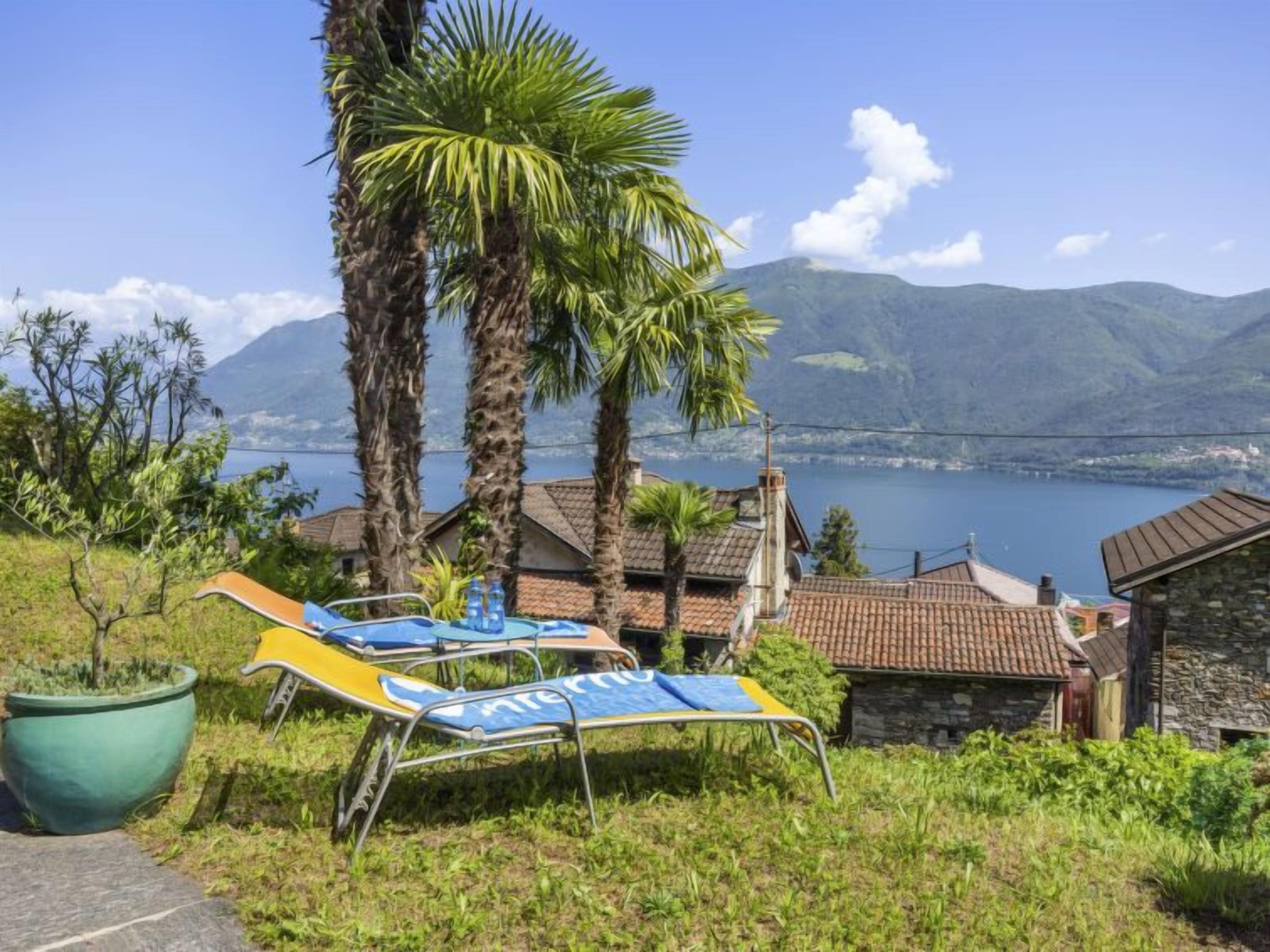 Vista de una propiedad frente al lago, con dos palmeras, un edificio de piedra y una fila de sillas reclinables azules y amarillas mirando hacia el agua.