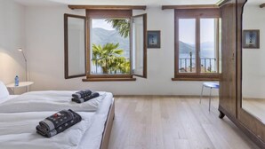 Un dormitorio con una cama grande, un armario, una mesita y una silla. Hay dos ventanas con vistas a montañas y un balcón con plantas.