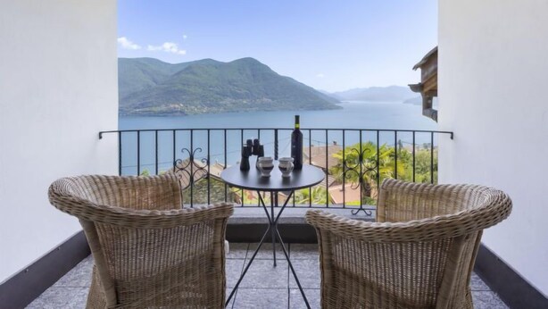 Un balcón con sillas de mimbre y una mesa, con vista a un lago y monta ñas.