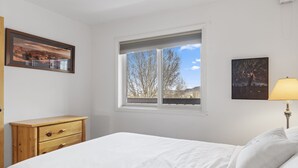 Une chambre à coucher avec un lit, une fenêtre donnant sur des arbres, une commode en bois et deux tableaux encadrés accrochés au mur.