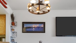 Un coin repas comprenant une table ronde en bois, six chaises en bois, un lustre, un tableau encadré au mur et un téléviseur à écran plat.