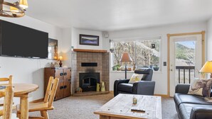 Un salon avec une cheminée, un téléviseur, une table à manger avec des chaises, un canapé et une vue sur les arbres par la fenêtre.