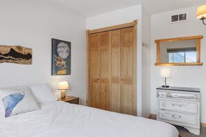 A bedroom with a bed, a wooden wardrobe, a dresser, a nightstand, a lamp, and wall art.