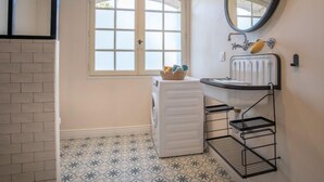 Un baño con suelo de baldosas blancas, una ventana con vidrio esmerilado, un radiador blanco, un espejo redondo y una estantería de metal.