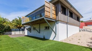 Une maison moderne à deux étages, dotée d’une grande baie vitrée à l’étage supérieur, d’une terrasse en bois et d’une pelouse bien entretenue.