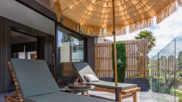 Un espace de détente extérieur moderne, comprenant deux fauteuils, une terrasse en bois et un grand parasol pour créer de l’ombre.