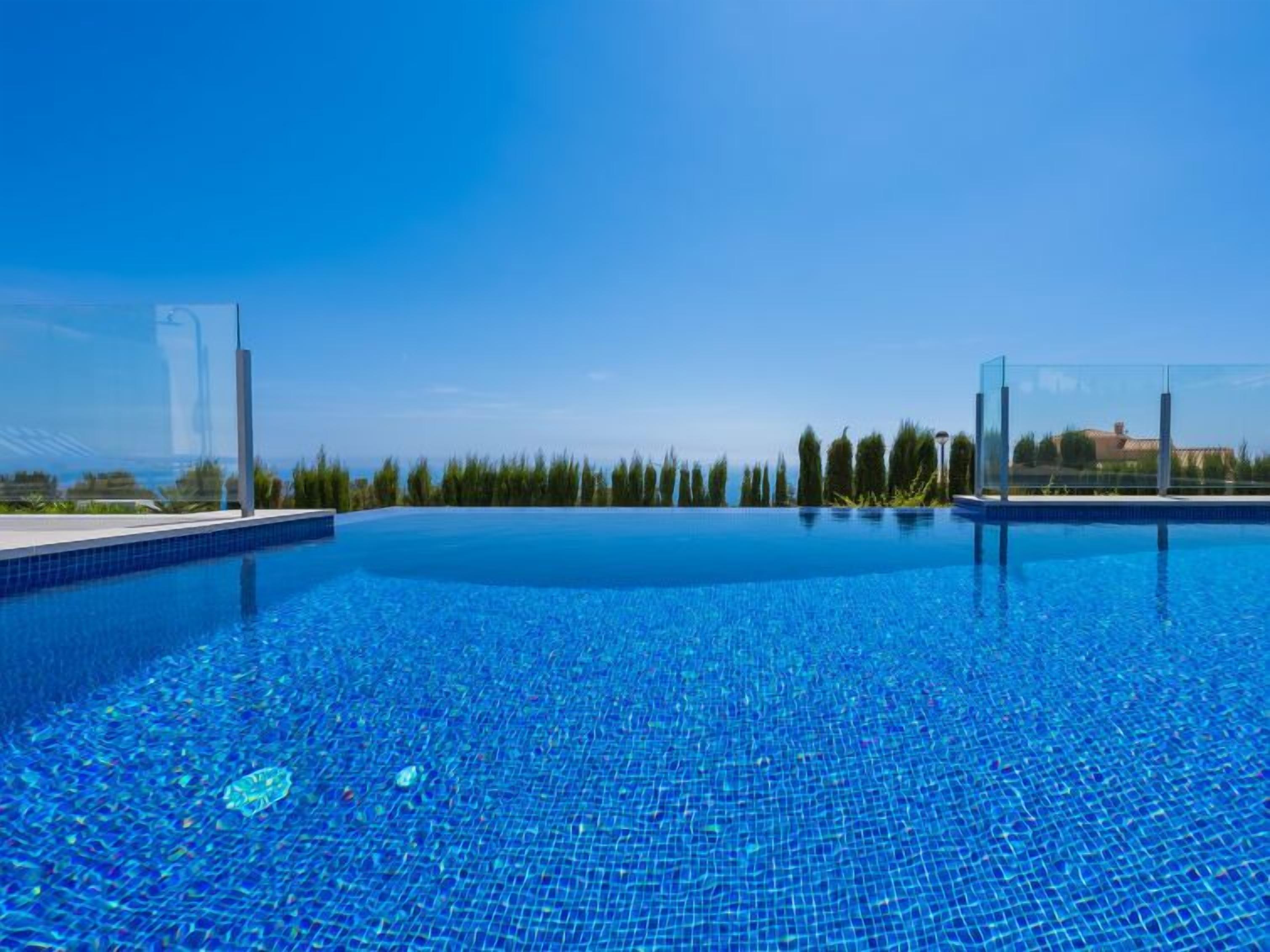 Uma piscina azul cristalina com borda azulejada, cercada por uma grade e árvores, sob um céu limpo.