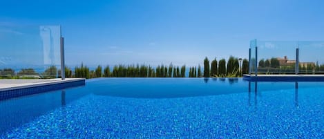 Uma piscina azul cristalina com borda azulejada, cercada por uma grade e árvores, sob um céu limpo.