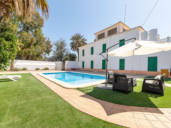 Une piscine agrémentée d’un parasol blanc, entourée de gazon synthétique et bordée d’un chemin pavé.
