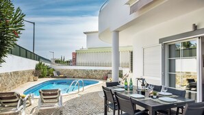 A poolside area with a table set for dining, a pool, and a view of a building in the background. - Beautiful detached Villa, 10 minutes walk from Porto Do Mos beach (Lagos)