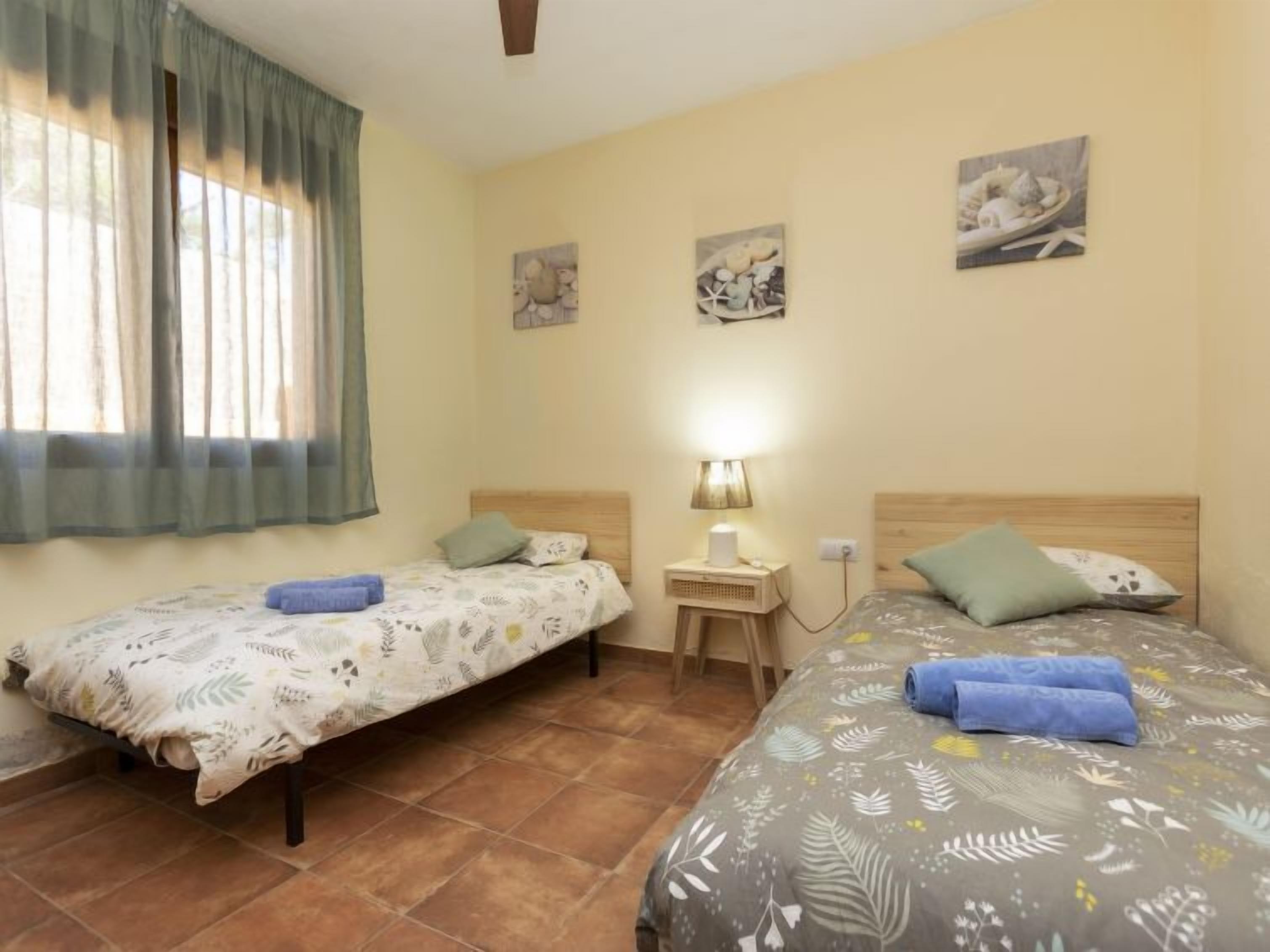A room with two beds, a small table with a lamp, and artwork on the walls.