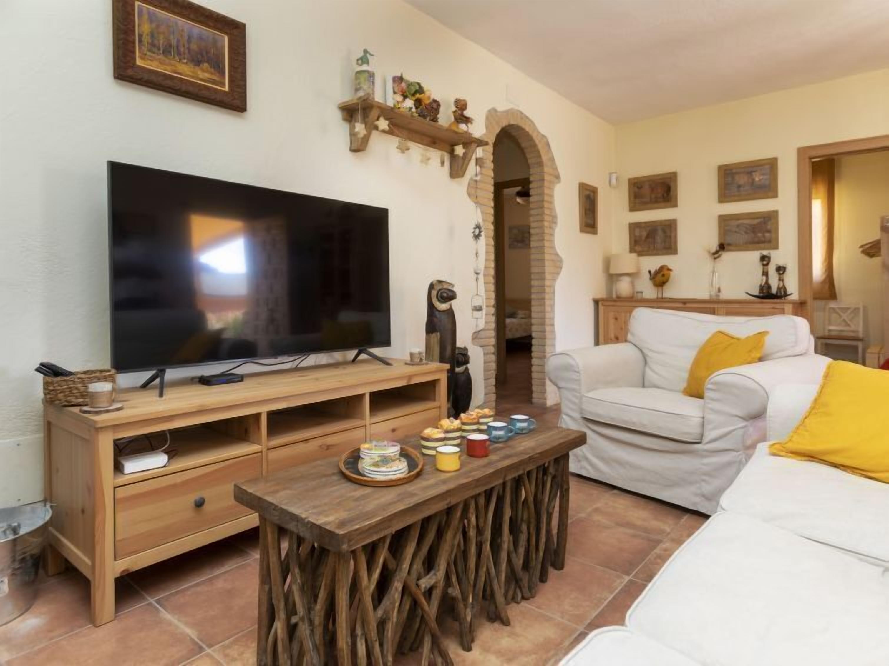 Un salon avec un grand téléviseur à écran plat posé sur un meuble en bois, un canapé blanc agrémenté d’un coussin jaune et une table basse en bois au style rustique.