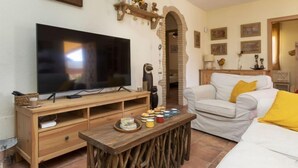 A living room with a large flat-screen TV on a wooden stand, a white sofa with a yellow pillow, and a rustic wooden coffee table. - Ca La Lou by Interhome (Sils)