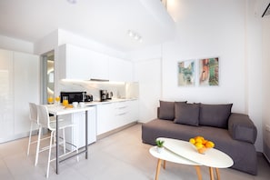 A modern kitchen with white cabinets, a dining area with white chairs, a gray sofa, and a small table with fruit.