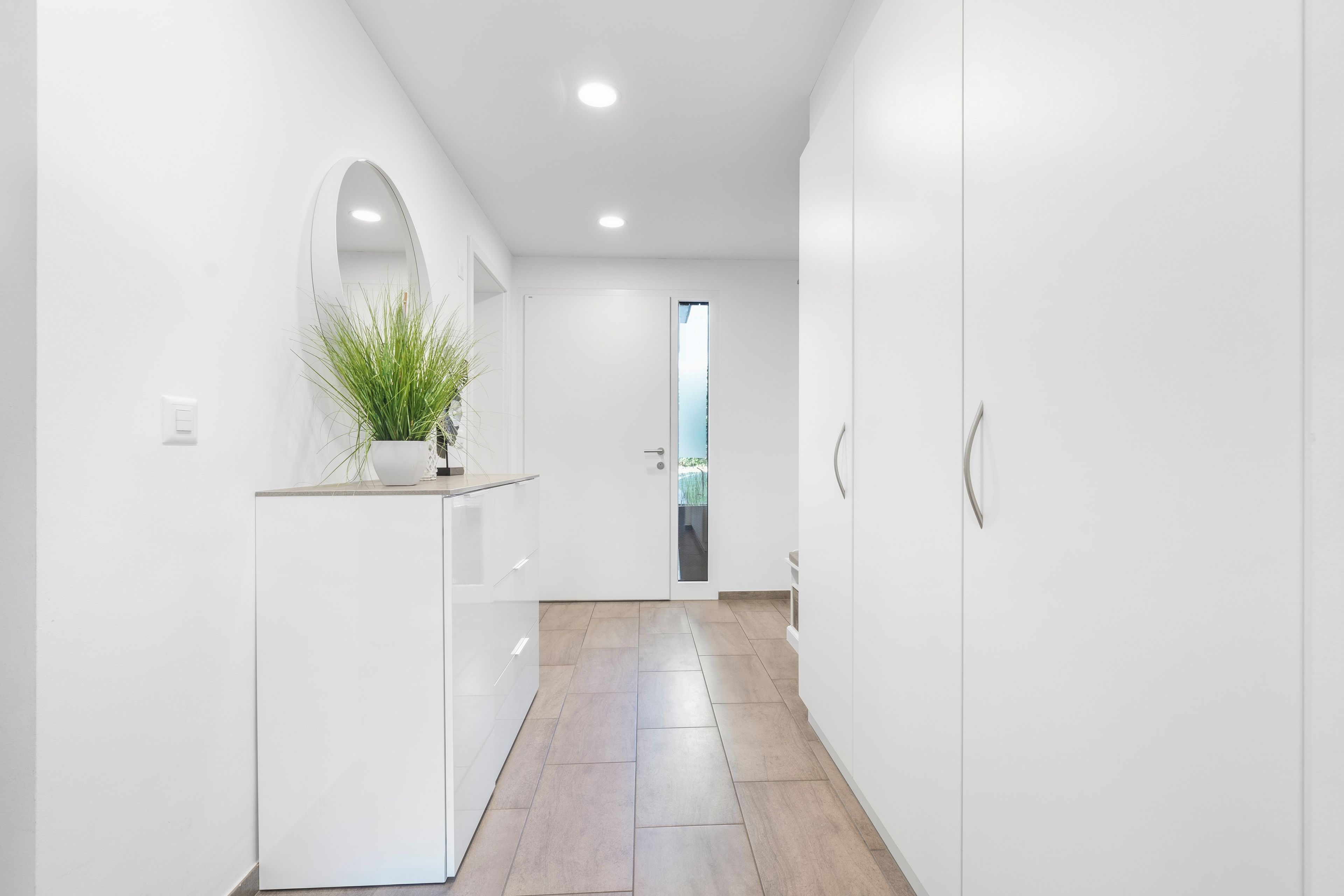 Un couloir intérieur moderne, aux murs blancs, agrémenté d’une plante en pot, avec une porte donnant sur une autre pièce et un sol carrelé.
