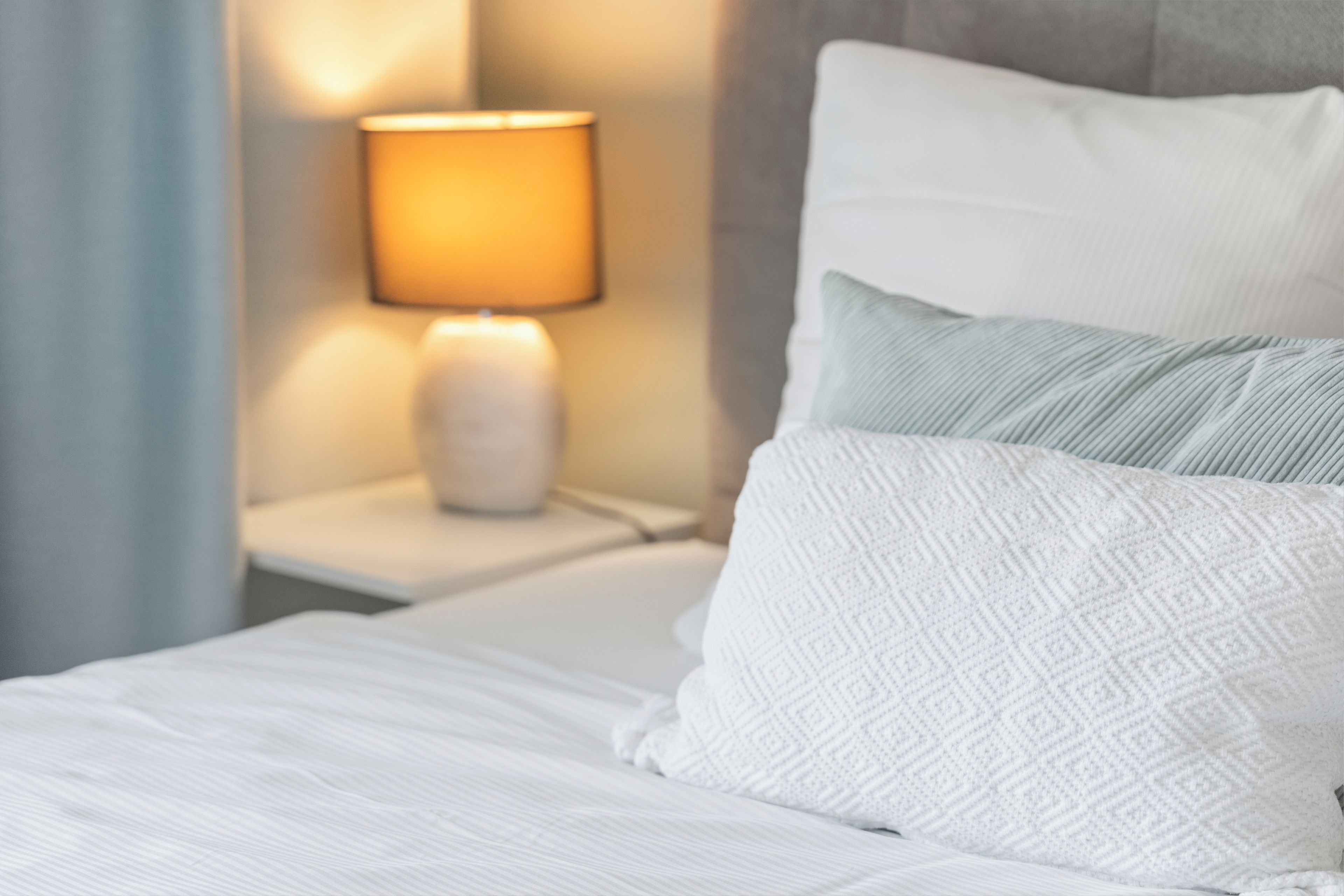 Ein ordentlich bezogenes Bett mit weißer Bettwäsche und Kissen, eine Nachttischlampe und ein Nachttisch.