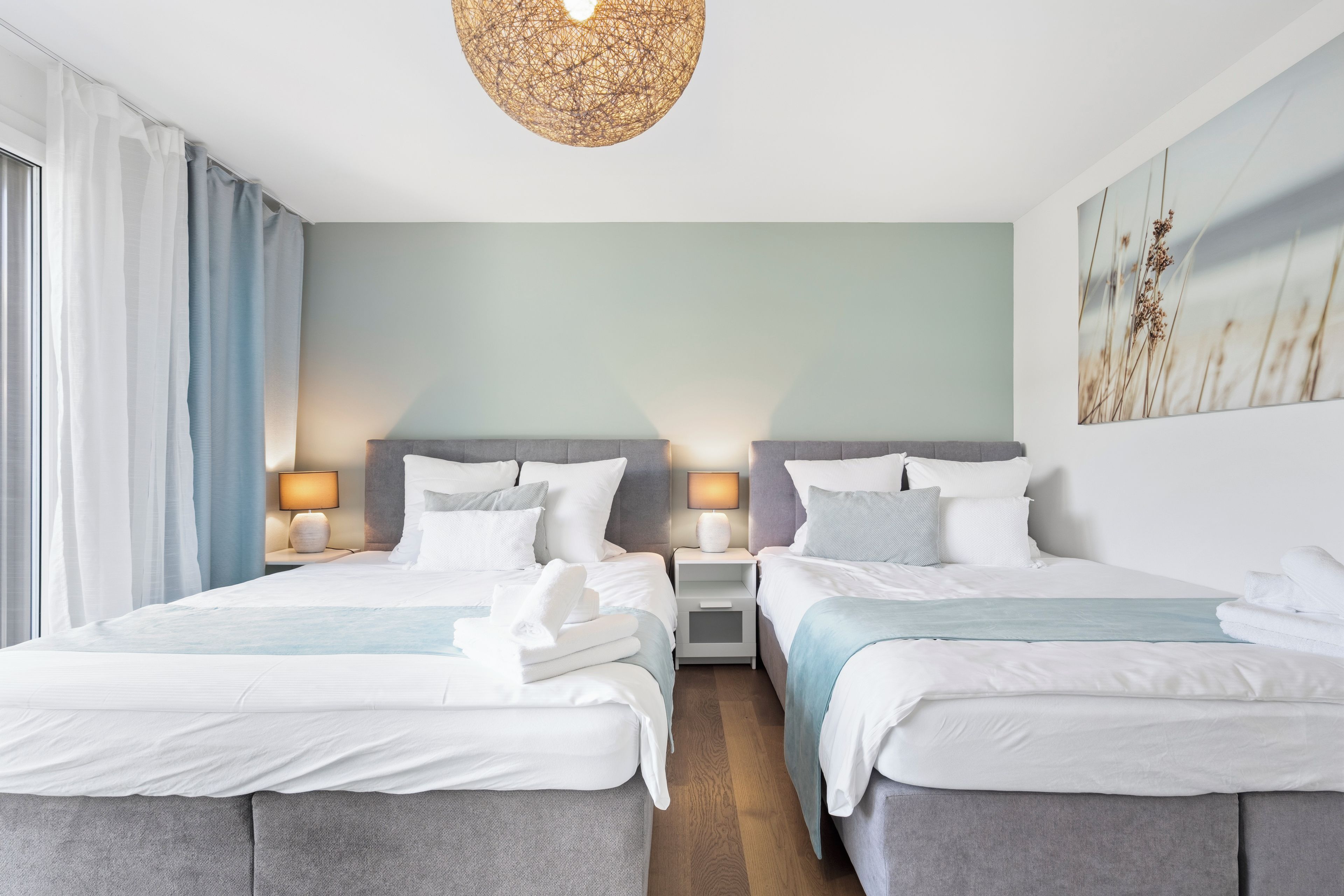 Ein Hotelzimmer mit zwei Betten, einem großen Fenster mit Vorhängen, einer Wandleuchte und einem Gemälde.