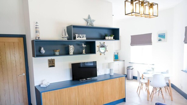 Un salon moderne avec un meuble en bois, un téléviseur à écran plat et un coin repas comprenant des chaises blanches et une table.