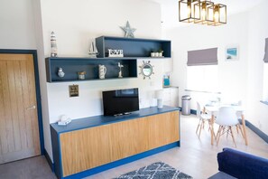 A modern living room with a wooden cabinet, a flat-screen TV, and a dining area with white chairs and a table. - Charming 1-bed Apartment in Bournemouth Near sea (Bournemouth)