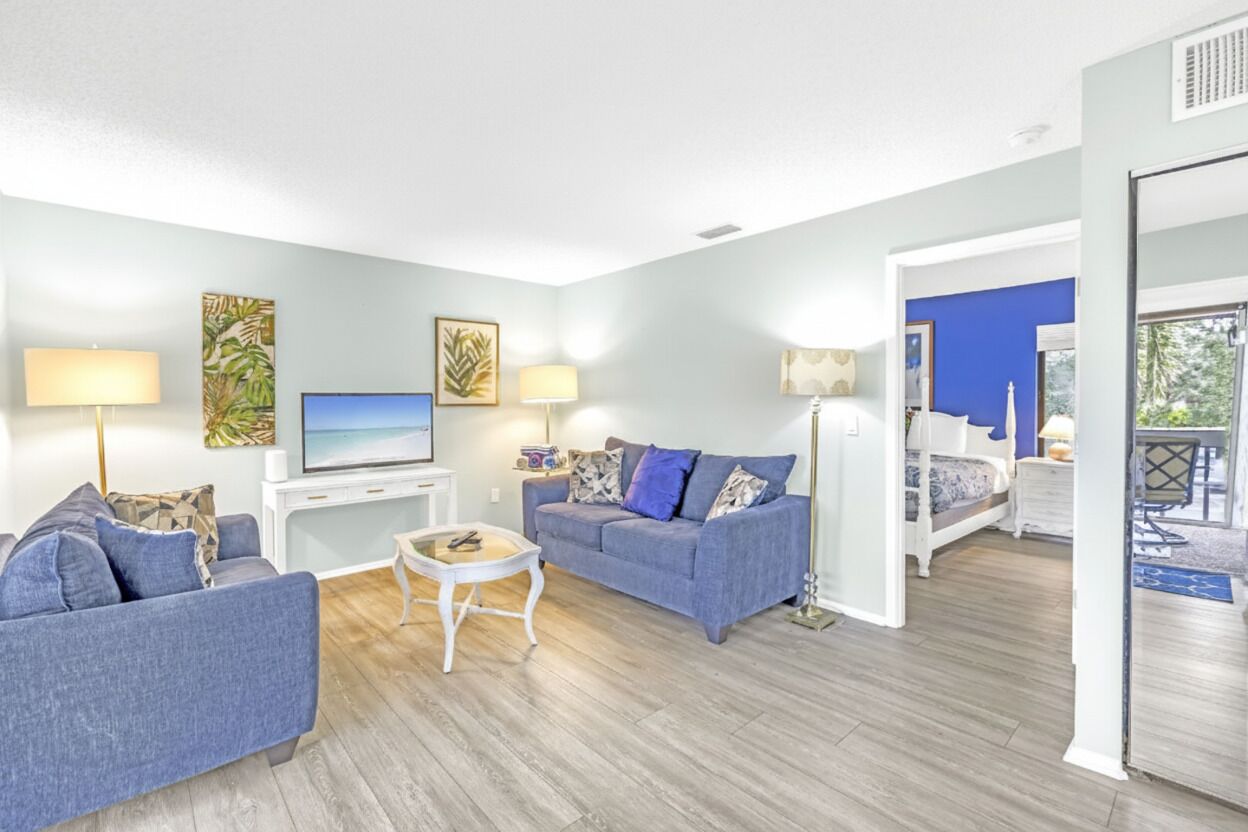 A modern living room with a grey sofa, a white coffee table, and a flat-screen TV. There is a blue accent wall and a balcony with a view.