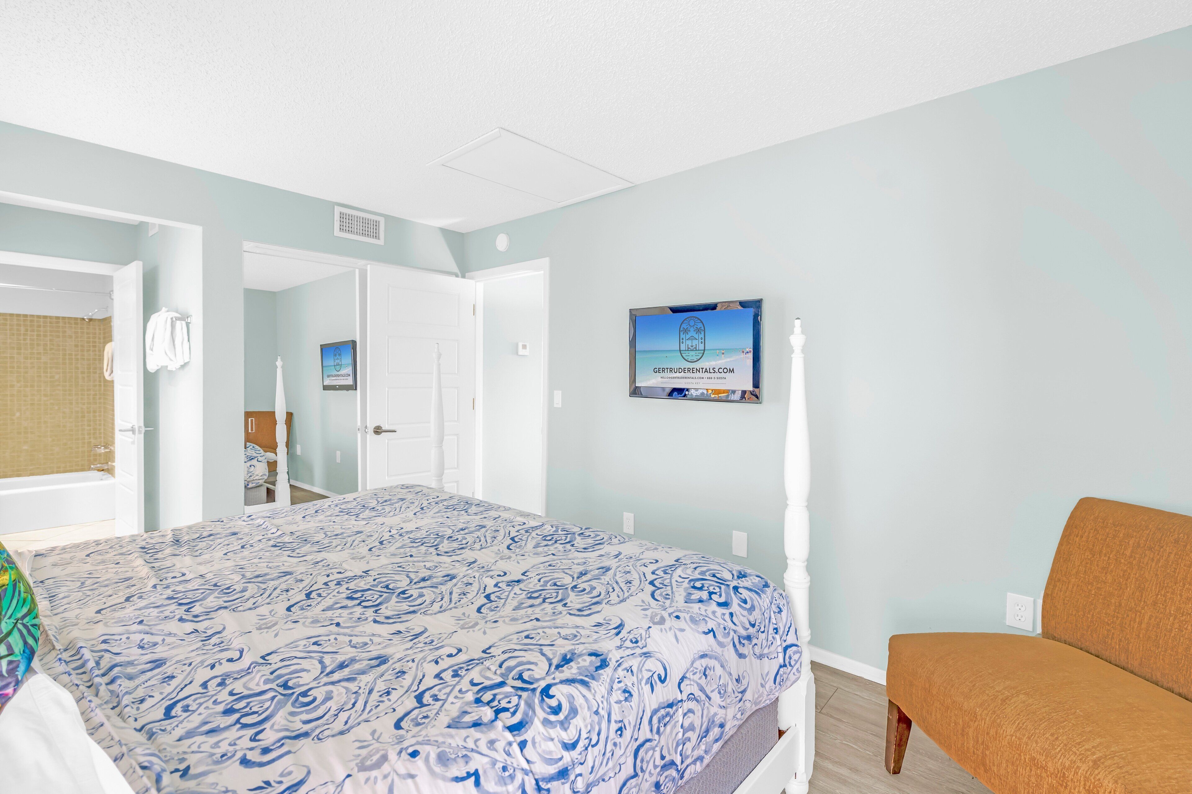 A bedroom with a bed, a chair, a television, and a bathroom visible through an open door.