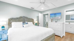 Une chambre avec un grand lit, un ventilateur de plafond, un téléviseur fixé au mur et une salle de bain visible à travers une porte ouverte.