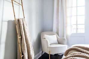 A room with a ladder leaning against a wall, a white armchair with a cushion, and a window with curtains.